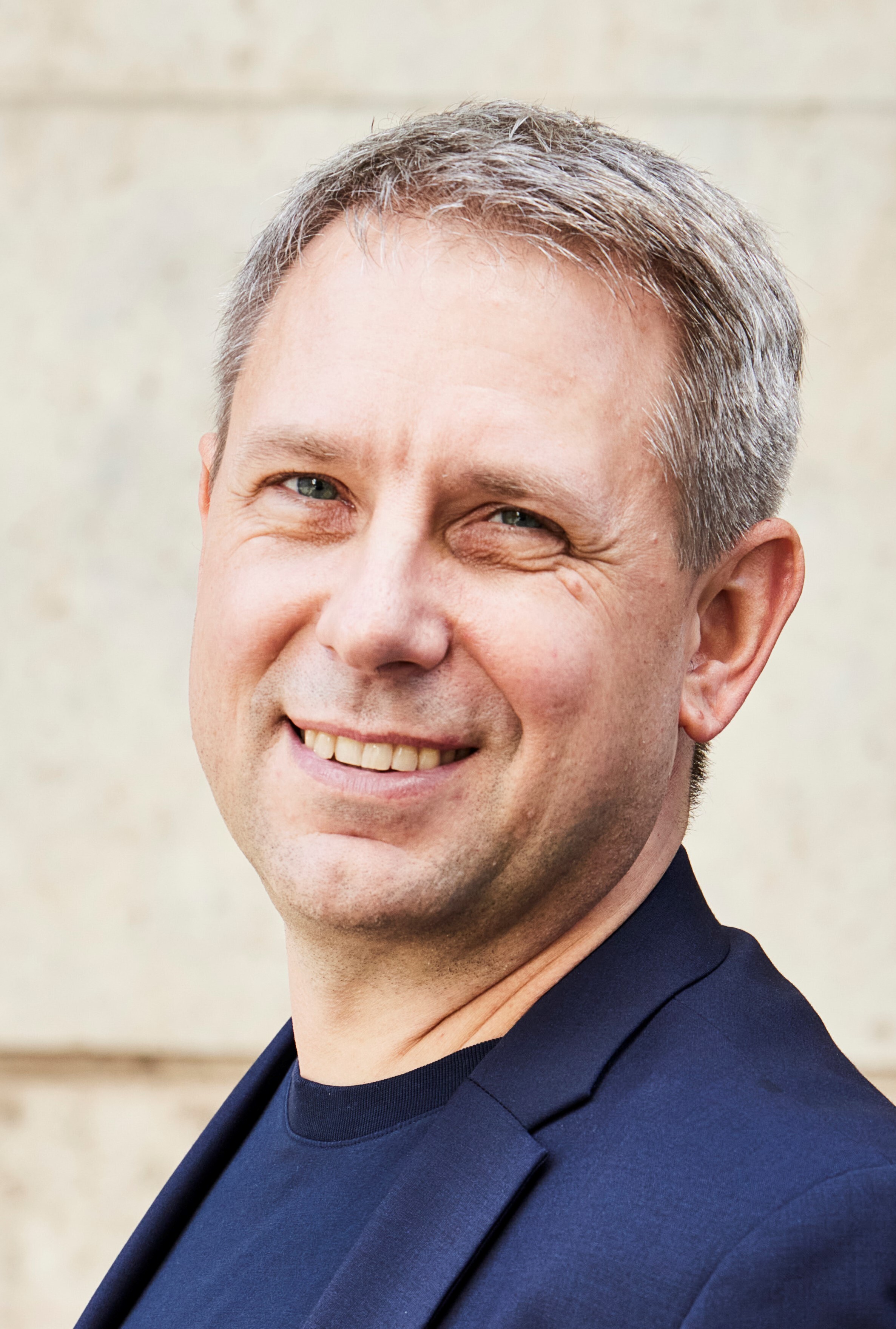 Portrait eines Mannes mit Namen Dr. Niels Pinkwart. Er hat graues Haar und trägt dunkelblauen Blazer und schwarzen Pullover vor heller Wand.