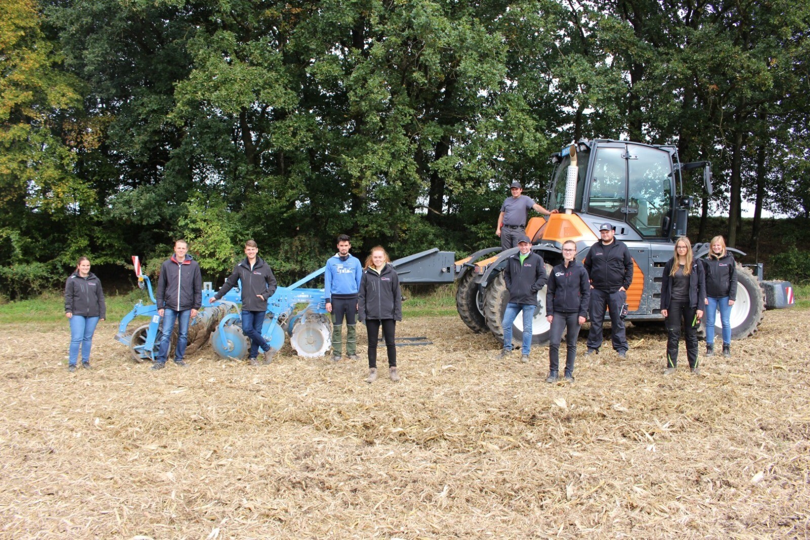 Eine Gruppe Studenten stehen vor einem Pflug und einem Traktor auf einem Feld