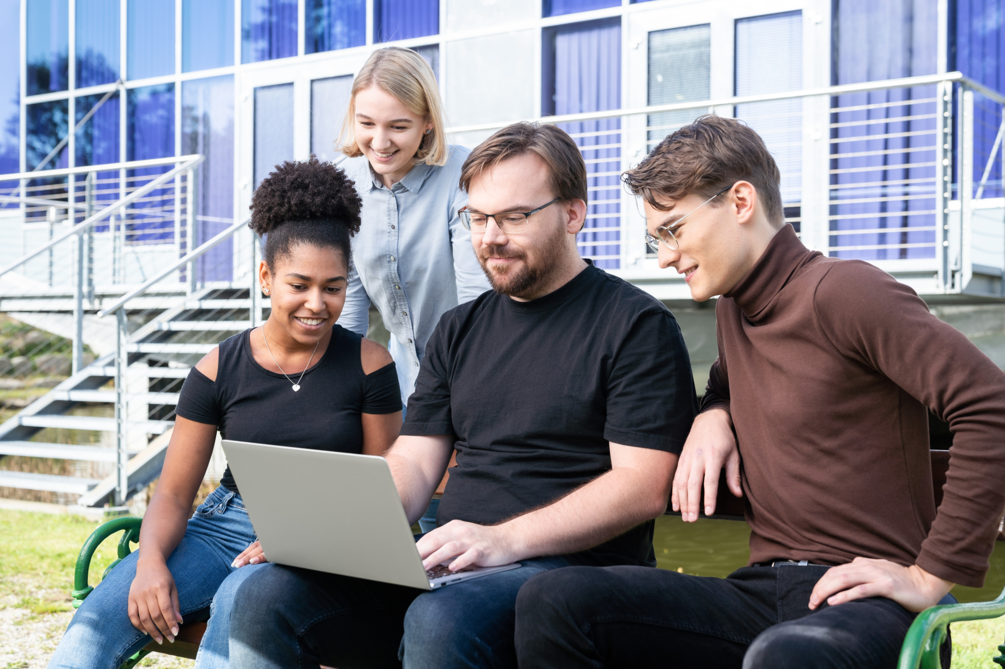 Vier Studenten sitzen auf einer Bank und schauen alle auf den Laptop des Studenten in der Mitte