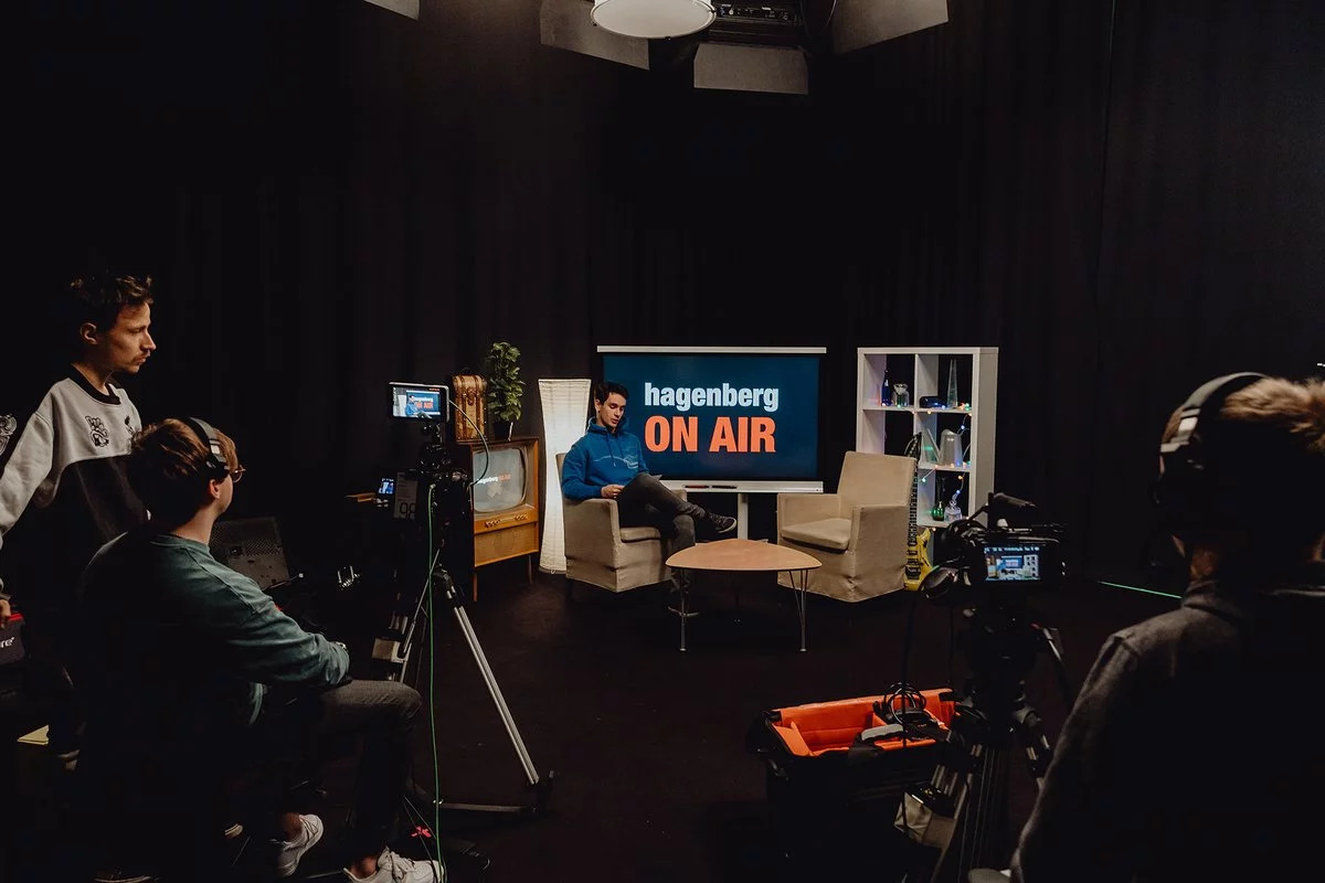 In einem dunklen Raum wird ein Interview gehalten. Ganz hinten ist eine kleine Leinwand und darauf geschrieben steht: Hagenberg on air