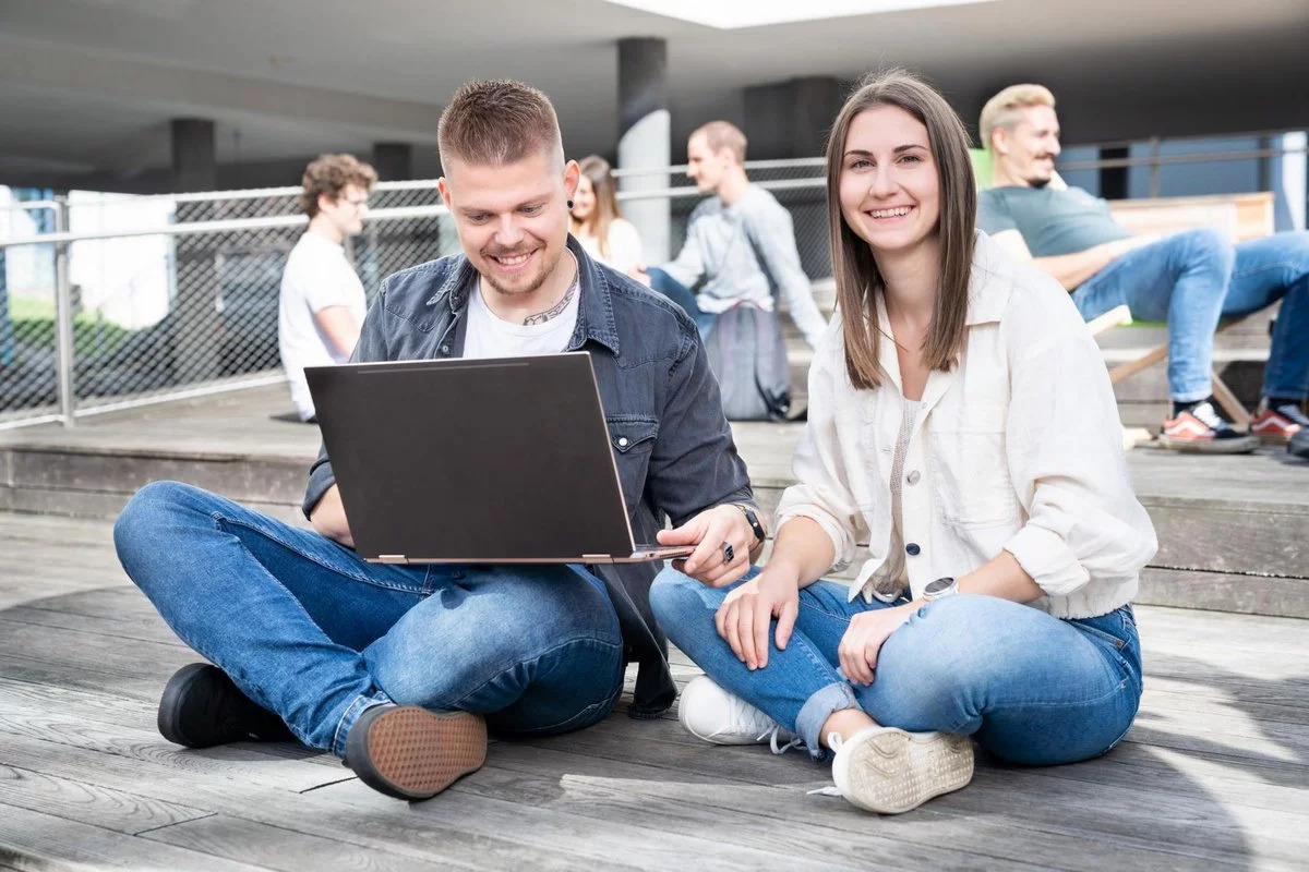 Eine Studentin und ein Student sitzen im Schneidersitz am Boden. Der Student hat einen Laptop auf dem Schoß.