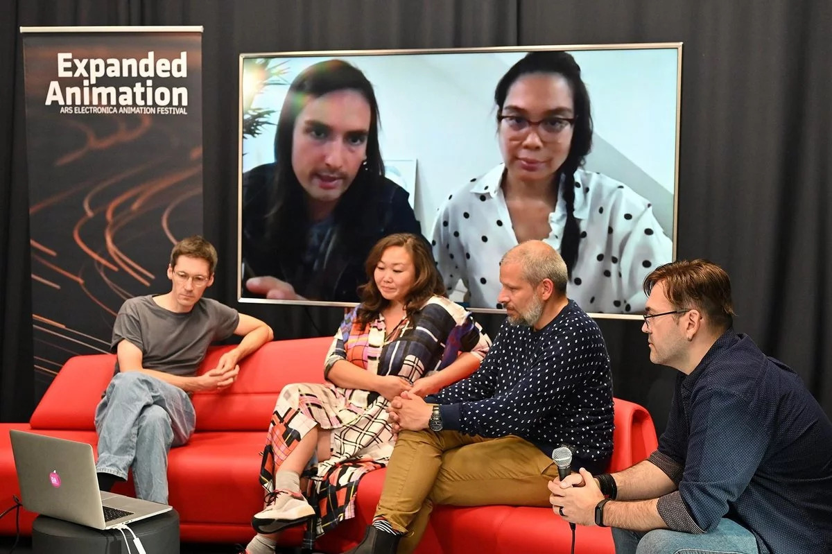 Zwei Männer und eine Frau sitzen auf einer roten Couch und diskutieren mit einem Mann, der neben ihnen auf einem Stuhl sitzt und ein Mikrofon in der Hand hält. Im Hintergrund sieht man auf einer Leinwand zwei weitere Personen.