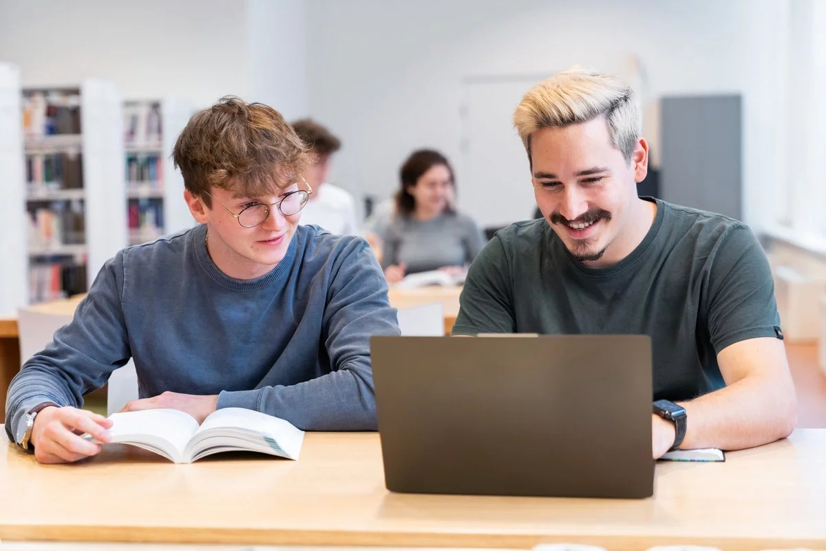 Zwei junge Männer sitzen zusammen an einem Tisch in einem Klassenzimmer. Einer der beiden liest ein Buch und der andere arbeitet an seinem Laptop.