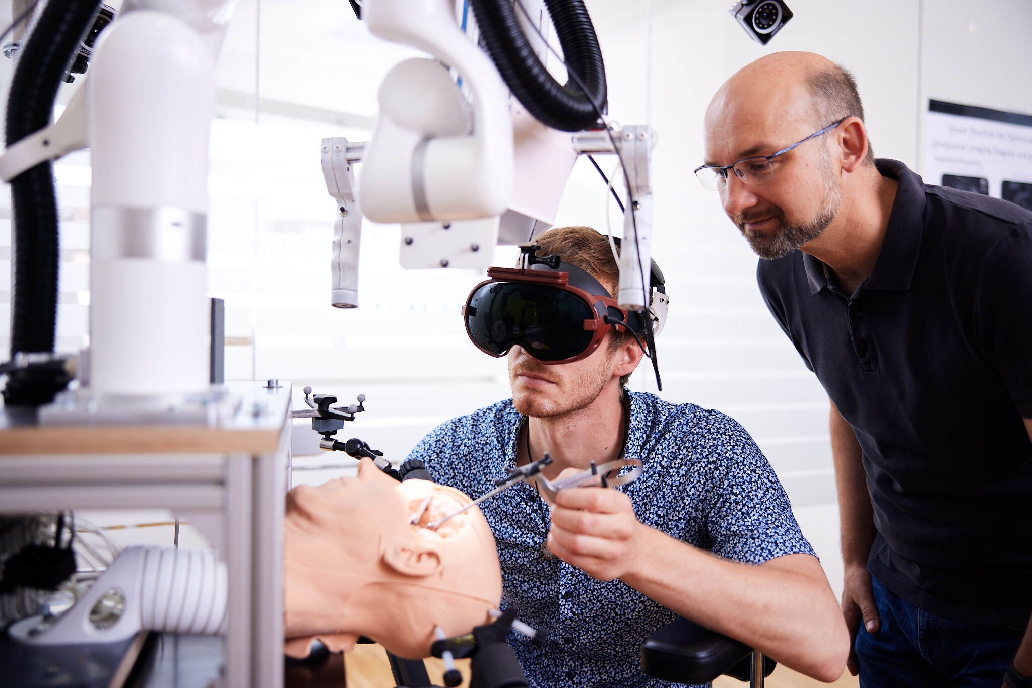 ein junger Student mit einem VR Headset operiert mit technischen Geräten an einem Test-Gehirn, während ein älterer Mann hinter ihm steht