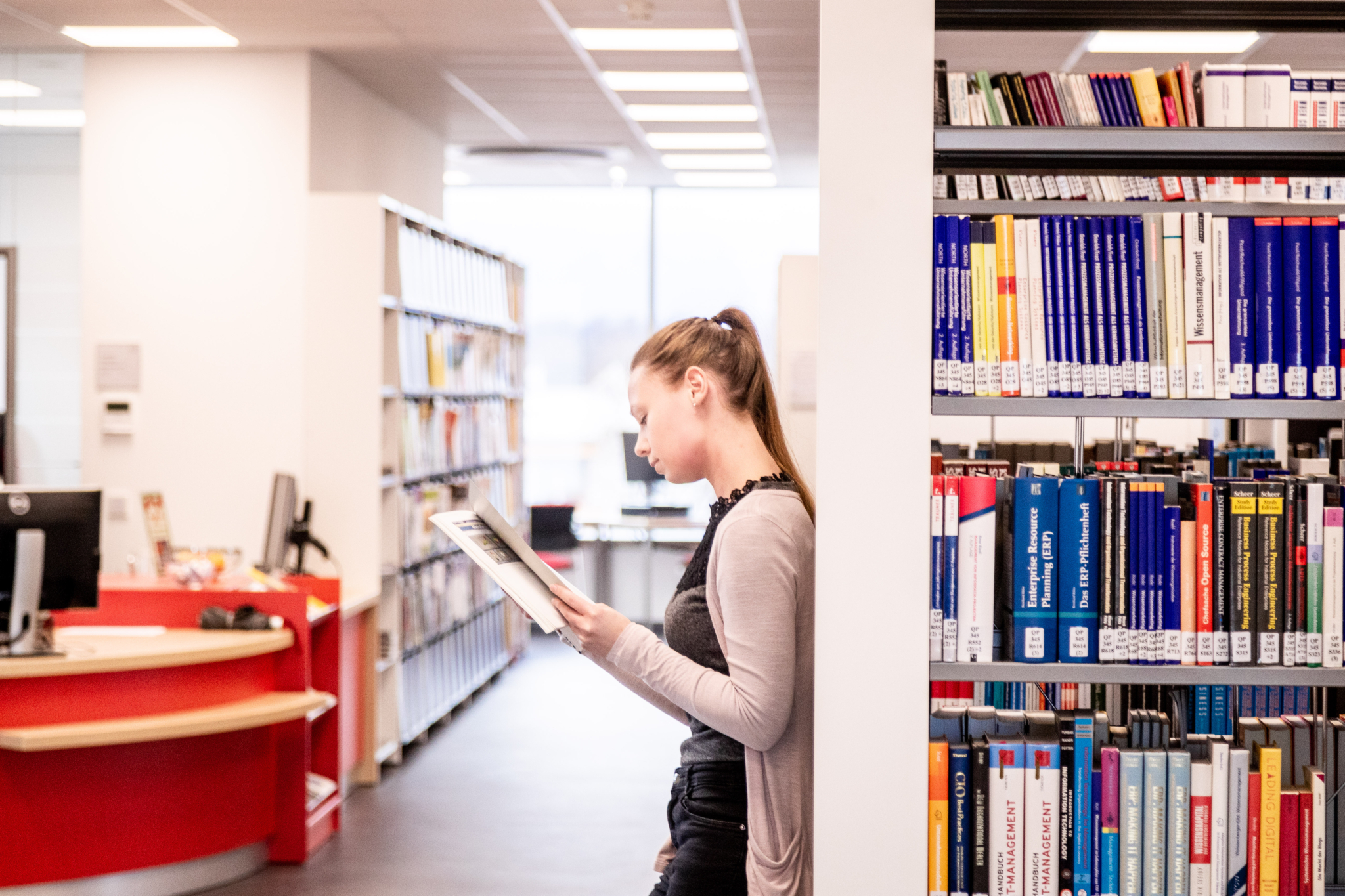 Eine weibliche Person lehnt an einem Bücherregal und liest ein Buch. Im Hintergrund sieht man die Bürotheke.